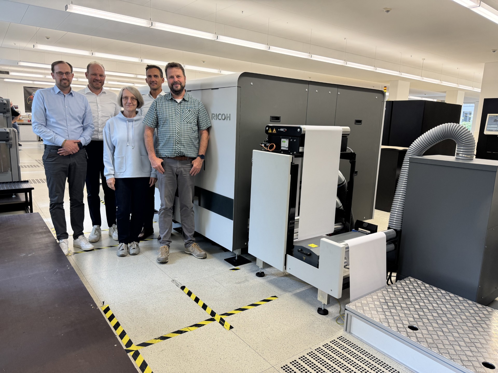 Group of professionals standing beside an industrial Ricoh printing machine in a modern production facility, showcasing advanced print technology, teamwork and operational excellence in a clean, well-lit workspace.