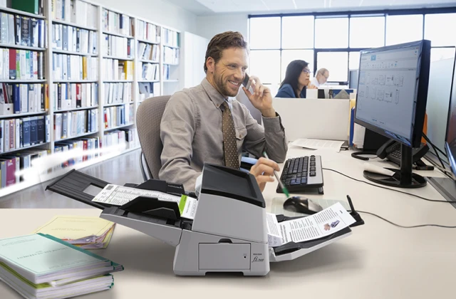 An office worker in a white-walled office with bookshelves in the background smiles while talking on a phone and using a desktop scanner. The scanner is actively feeding documents. Other blurred office workers and computer monitors are visible in the background.